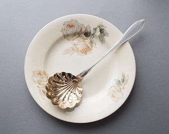 May include: A white porcelain plate with a floral design featuring white roses and green leaves. A silver spoon with a gold, scalloped, perforated bowl sits on the plate.