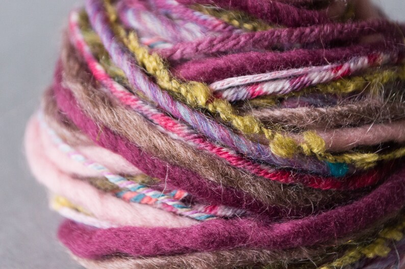 May include: A close-up of a ball of yarn with multiple colors, including pink, brown, red, green, and purple. The yarn is soft and fluffy, and it appears to be hand-spun.