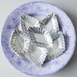 Può includere: Sei piccoli stampini argentati per tartellette scanalate disposti su un piatto a motivi blu e bianchi. Il piatto ha un bordo decorativo e un disegno centrale. Gli stampini sono vuoti e lucidi.