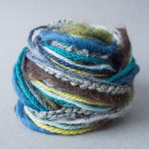 Puede incluir: Un ovillo de lana de varios colores: azul, verde, amarillo y marrón. La lana está hecha de diferentes texturas, incluyendo pelusa y lisa.