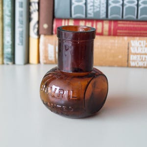 May include: A vintage brown glass bottle with a rounded base and cylindrical neck. The bottle has the embossed text "BOVRIL LIMITED" on its surface. The bottle is displayed in front of a collection of old books.