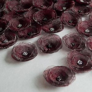 May include: A collection of burgundy fabric flower embellishments with silver and black bead centers. The flowers are arranged on a white surface, creating a textured and decorative display. These could be used for crafts or embellishments.