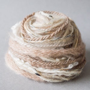 Könnte beinhalten: Ein Knäuel Garn in Beige-, Weiß- und Brauntönen. Das Garn besteht aus verschiedenen Fasern, darunter Wolle, Seide und Baumwolle. Das Garn ist weich und flauschig und eignet sich perfekt zum Stricken oder Häkeln.