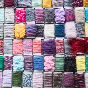 Puede incluir: Una colección de muestras de hilo en varios colores y texturas, incluyendo hilos peludos, esponjosos y lisos. El hilo está dispuesto en filas sobre una superficie blanca.