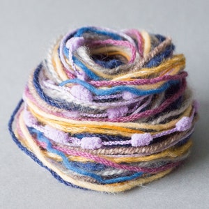 Puede incluir: Un ovillo de hilo de varios colores, incluyendo amarillo, azul, rosa y gris. El hilo tiene pequeños pompones esponjosos en todo.