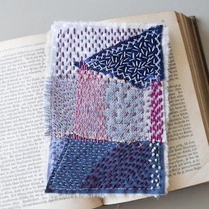 Pode incluir: Uma obra de arte têxtil feita à mão com várias peças de tecido em tons de azul, roxo e branco, costuradas juntas. A obra de arte é colocada em um livro aberto com páginas envelhecidas, mostrando uma mistura de texturas e cores.