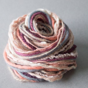 Puede incluir: Un ovillo de hilo en tonos de rosa, melocotón, gris y blanco. El hilo está enrollado en forma de bola.