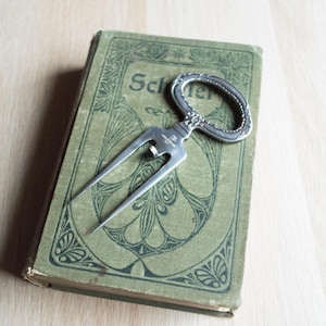 Puede incluir: Un abridor de botellas plateado antiguo descansa sobre un libro verde vintage con la palabra "Schler" visible. El libro tiene un diseño ornamentado y está ligeramente desgastado. El abridor de botellas tiene un mango decorativo y un diseño en forma de tenedor.