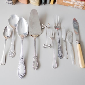 Puede incluir: Un juego de cubiertos de plata, que incluye una cuchara de servir, un cuchillo para pastel, un cuchillo para mantequilla, una cuchara pequeña, un tenedor y dos tenedores para cenar. Los cubiertos están dispuestos sobre una superficie blanca.