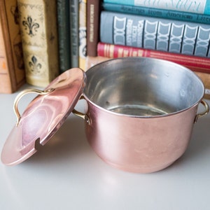 Puede incluir: Una olla de cobre con tapa y asas de latón. La olla está vacía y la tapa está quitada. La olla está sobre una superficie blanca.