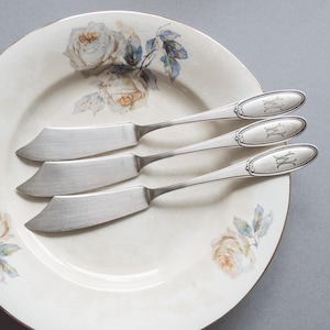 Op de afbeelding: Drie zilveren botermesjes met sierlijke handvatten en gegraveerde initialen "WW" op een wit bord met een bloemenmotief.