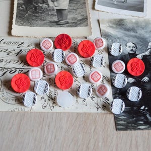 May include: A selection of vintage buttons in red, white, and black, arranged on a postcard and old photographs. The buttons display diverse geometric and floral patterns, evoking a sense of nostalgia.