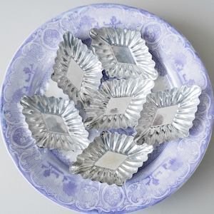 Puede incluir: Seis moldes para hornear de metal de color plateado con un diseño acanalado, dispuestos en un plato con un patrón azul y blanco. Los moldes tienen forma de diamantes con bordes redondeados.