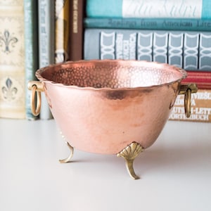 May include: A hammered copper bowl with brass handles and feet. The bowl has a slightly scalloped rim and a textured surface.