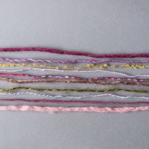 May include: A collection of colorful yarn strands in various shades of pink, purple, brown, and gold. The yarn is arranged in rows on a gray background.