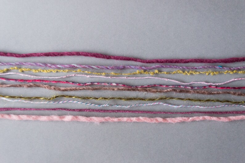 May include: A collection of colorful yarn strands in various shades of pink, purple, brown, and gold. The yarn is arranged in rows on a gray background.