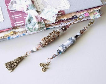 May include: Two handmade journal charms with decorative beads and tassels. One charm features a bicycle charm. The journal is gray with floral fabric accents and a fabric tie.