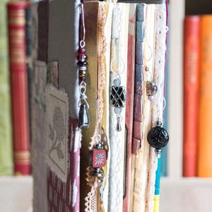 May include: Close-up of a handmade journal with several decorative beaded bookmarks. The journal has a fabric cover with a floral design and lace trim. The bookmarks feature various beads and charms in silver and dark colors.