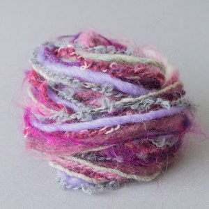 Puede incluir: Un ovillo de lana con hilos rosas, morados y grises. La lana es suave y esponjosa, y sería perfecta para tejer o hacer ganchillo.
