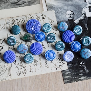 May include: A selection of vintage buttons in shades of blue and teal, arranged on antique postcards and beside a black and white photograph. The buttons display diverse designs, including swirls, woven textures, and floral patterns.