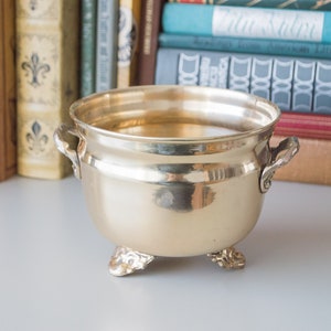 May include: A gold-toned brass planter with three decorative feet and two handles. The planter is sitting on a white surface in front of a bookshelf.
