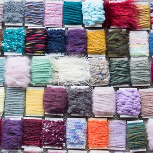 Puede incluir: Una colección de muestras de hilo de colores variados y texturas diferentes, incluyendo peludo, esponjoso y liso. El hilo está dispuesto en filas sobre una superficie blanca.