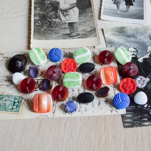 May include: A selection of vintage buttons in diverse colours and forms, such as red, orange, green, blue, and black. The buttons are displayed on a vintage postcard, alongside antique photographs.