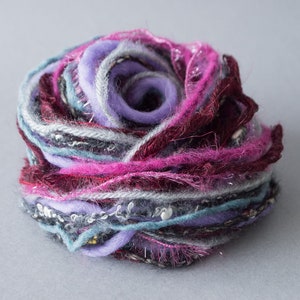 Puede incluir: Una bola de hilo hecha a mano, multicolor, en tonos morado, rosa, gris y turquesa. El hilo está bien enrollado, creando una forma texturizada similar a una flor. La bola de hilo está sobre un fondo gris liso.