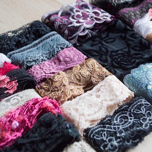 May include: A selection of lace trims in diverse colours, such as black, white, pink, and blue. The lace displays floral and geometric patterns, with some having beaded details. The trims are arranged in neat rows, highlighting their textures.