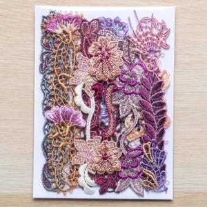 May include: A greeting card featuring a detailed floral lace design. The card showcases a variety of embroidered flowers and leaves in shades of purple, pink, and gold. The intricate lace is set against a white background.
