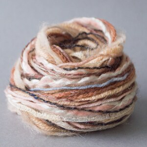 Può includere: Un gomitolo di filato con una miscela di fili beige, marrone, rosa e blu. Il filato è avvolto in modo lasco e ha una consistenza soffice.