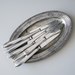 May include: An oval, antique silver serving tray holds several silver knives. The knives have ornate handles and are arranged in a fan shape. The tray has a decorative border and is tarnished.