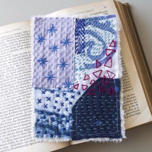 Può includere: Un'opera d'arte tessile con varie toppe di tessuto blu e viola. Le toppe presentano stelle ricamate, vortici e forme geometriche. L'opera d'arte è posta su un libro aperto con pagine invecchiate.