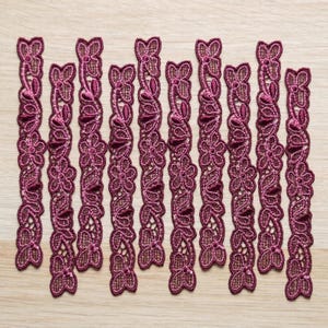 May include: A collection of burgundy lace trim pieces arranged on a light wood surface. Each piece features a floral and leaf design with intricate detailing. The trim is a deep, rich burgundy color, perfect for sewing and craft projects.