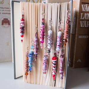 Puede incluir: Una colección de 10 marcapáginas de tela y cuentas de colores que cuelgan de un libro. Los marcapáginas están hechos con diferentes colores de tela y cuentas, incluyendo rojo, rosa, azul y marrón.