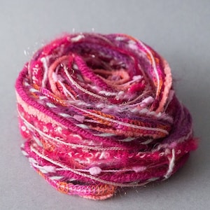 Puede incluir: Un ovillo de hilo en tonos rosa, naranja y blanco. El hilo está hecho de una variedad de fibras, incluyendo lana, seda y algodón. Es un hilo suave y esponjoso que sería perfecto para tejer o hacer ganchillo.