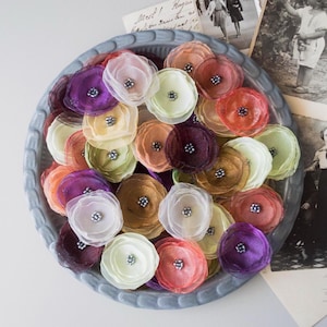 May include: A variety of fabric flower embellishments in a grey, scalloped-edge bowl. The flowers are in shades of purple, pink, orange, yellow, and green, each with a silver bead centre. Vintage photographs are in the background.