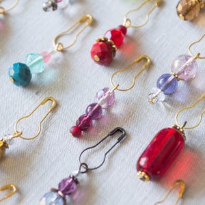 May include: A collection of colourful glass beads strung on gold safety pins. The beads are various shapes and sizes, including round, oval, and teardrop. The safety pins are open and ready to be used.