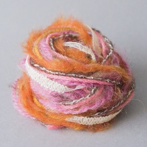 Puede incluir: Una bola de hilo con hebras rosadas, naranjas y marrones. El hilo está envuelto en una cinta blanca.