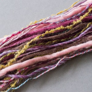 May include: A close-up of a bundle of yarn in various shades of pink, purple, brown, and gold. The yarn is soft and fluffy, and it appears to be hand-spun.