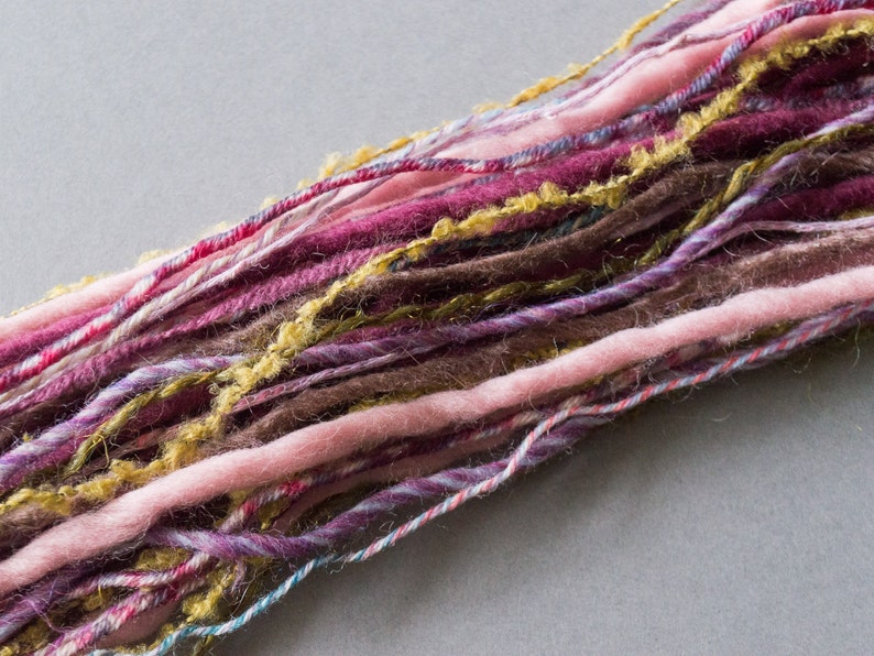 May include: A close-up of a bundle of yarn in various shades of pink, purple, brown, and gold. The yarn is soft and fluffy, and it appears to be hand-spun.