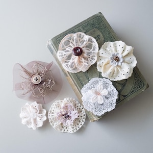May include: A selection of handmade fabric flower embellishments in various styles and colours. The flowers include lace, tulle, and other delicate materials, with button and bead centres. Some are placed on a vintage book.