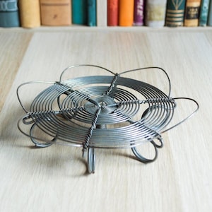 May include: A silver-colored, wire trivet with a spiral design and four curved feet. The trivet is made of thin metal wire and is designed to protect surfaces from hot cookware. The trivet is on a light wood surface.