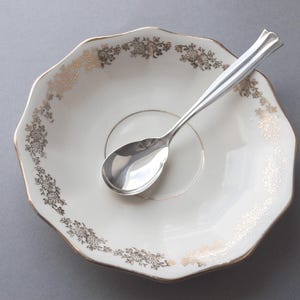 May include: A white scalloped saucer featuring a delicate gold floral pattern along the rim, paired with a polished silver-toned metal spoon resting in the centre. The saucer has a circular indentation and a thin gold line accenting the inner ring.