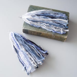 May include: Two blue and white yarn tassels with a black ribbon and white lace trim. The tassels are hanging from a silver ring.