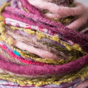 May include: A close-up of a ball of yarn with multiple colors, including pink, purple, green, and brown. The yarn is textured and has a fuzzy appearance.