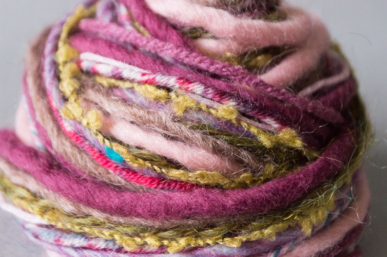 May include: A close-up of a ball of yarn with multiple colors, including pink, purple, green, and brown. The yarn is textured and has a fuzzy appearance.