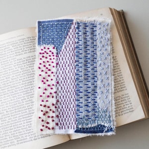 Può includere: Un'opera d'arte tessile fatta a mano con un design patchwork. Presenta sezioni di tessuto bianco, blu e viola, cucite in modo intricato con vari motivi. Il pezzo è appoggiato su un libro aperto, mostrando la consistenza e l'artigianato.
