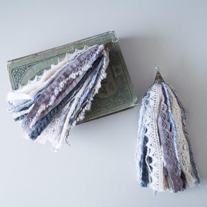May include: Two handmade fabric tassels in shades of white, gray, and blue. The tassels are made from a variety of fabrics, including lace, yarn, and ribbon. They are attached to silver metal rings.