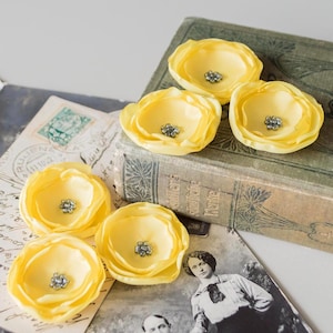 May include: Six yellow fabric flower embellishments with silver rhinestone centres. The flowers are arranged on vintage ephemera, including an old book and postcards. The flowers have a soft, ruffled petal design.